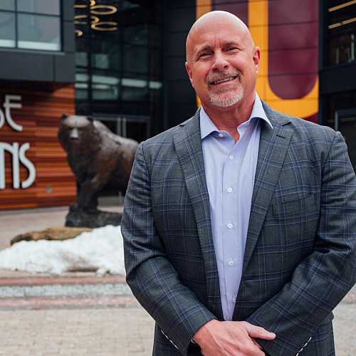 Outdoor photo of man in blazer in foreground with a bear statue in the background.