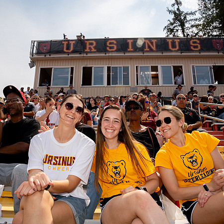 Group of three college students sitting attending a football game in Ursinus College merchandise.